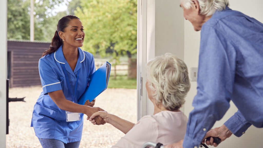 Senior Couple With Woman In Wheelchair Greeting Nurse Or Care Worker Making Home Visit At Door