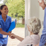 Senior Couple With Woman In Wheelchair Greeting Nurse Or Care Worker Making Home Visit At Door