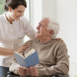 Photo of elderly man reading book and his personal home assistence