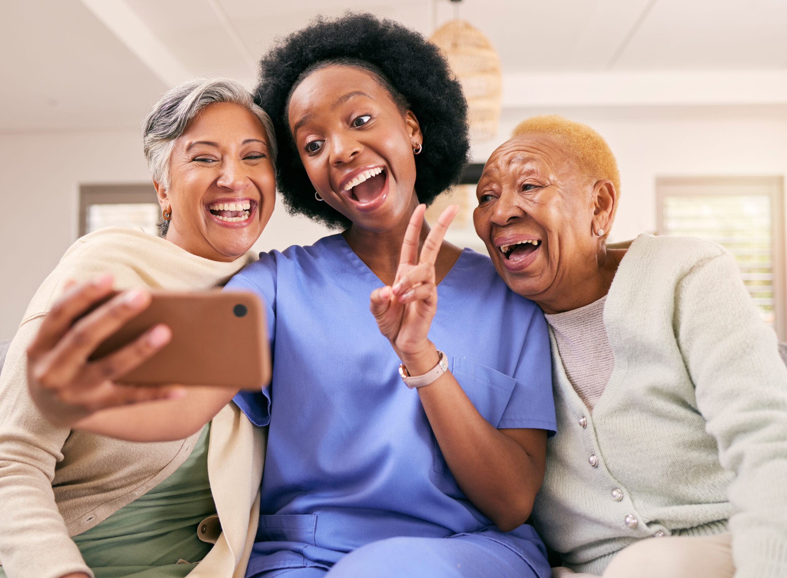 caregivers philadelphia pa Smiling female caregiver in blue scrubs taking a joyful selfie with two senior women, showing a peace sign. They are sitting together in a cozy home setting, sharing a happy moment.