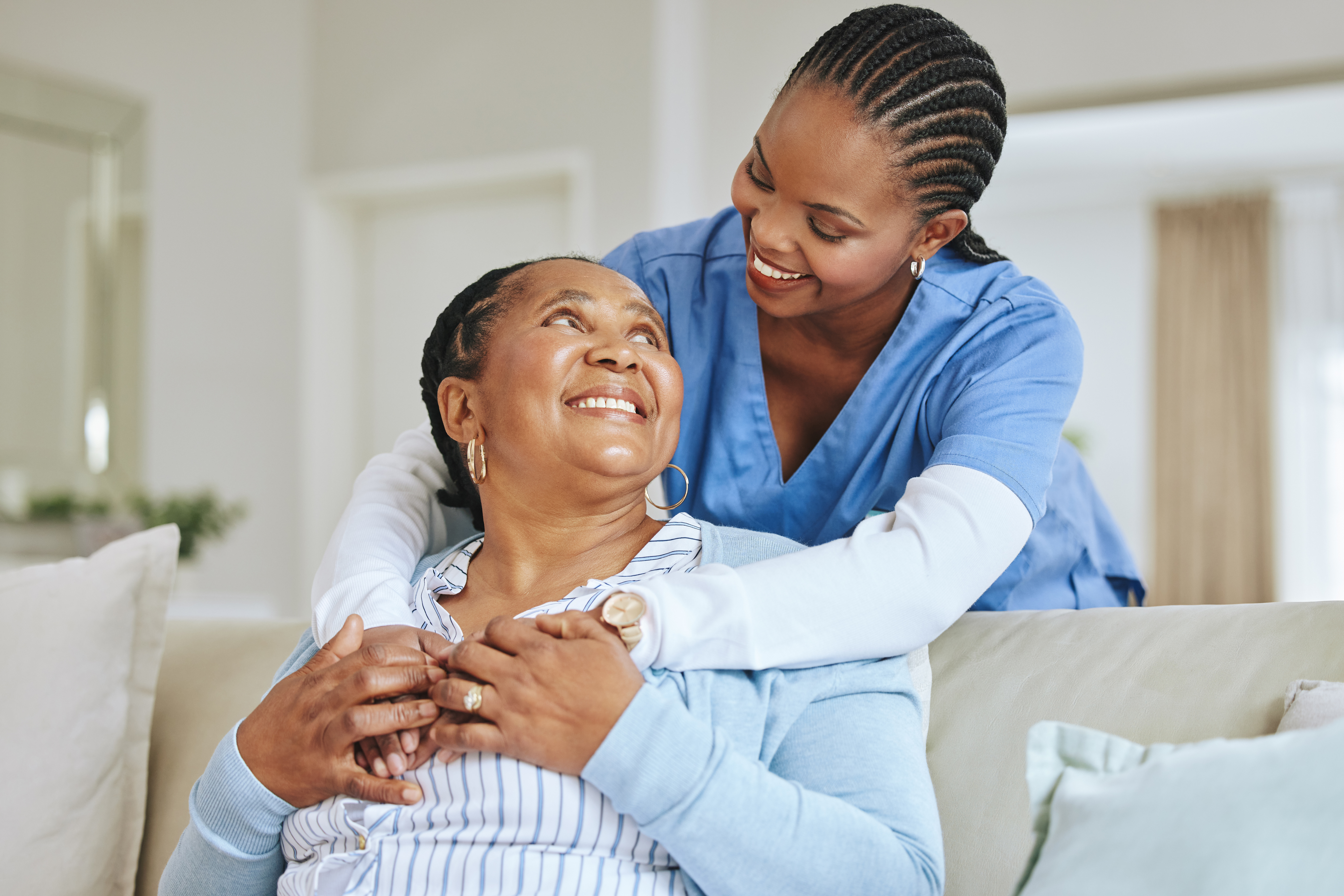 Smiling female caregiver in blue scrubs embracing a happy senior woman in a warm and caring home environment. They share a moment of connection and trust, symbolizing compassionate home care.