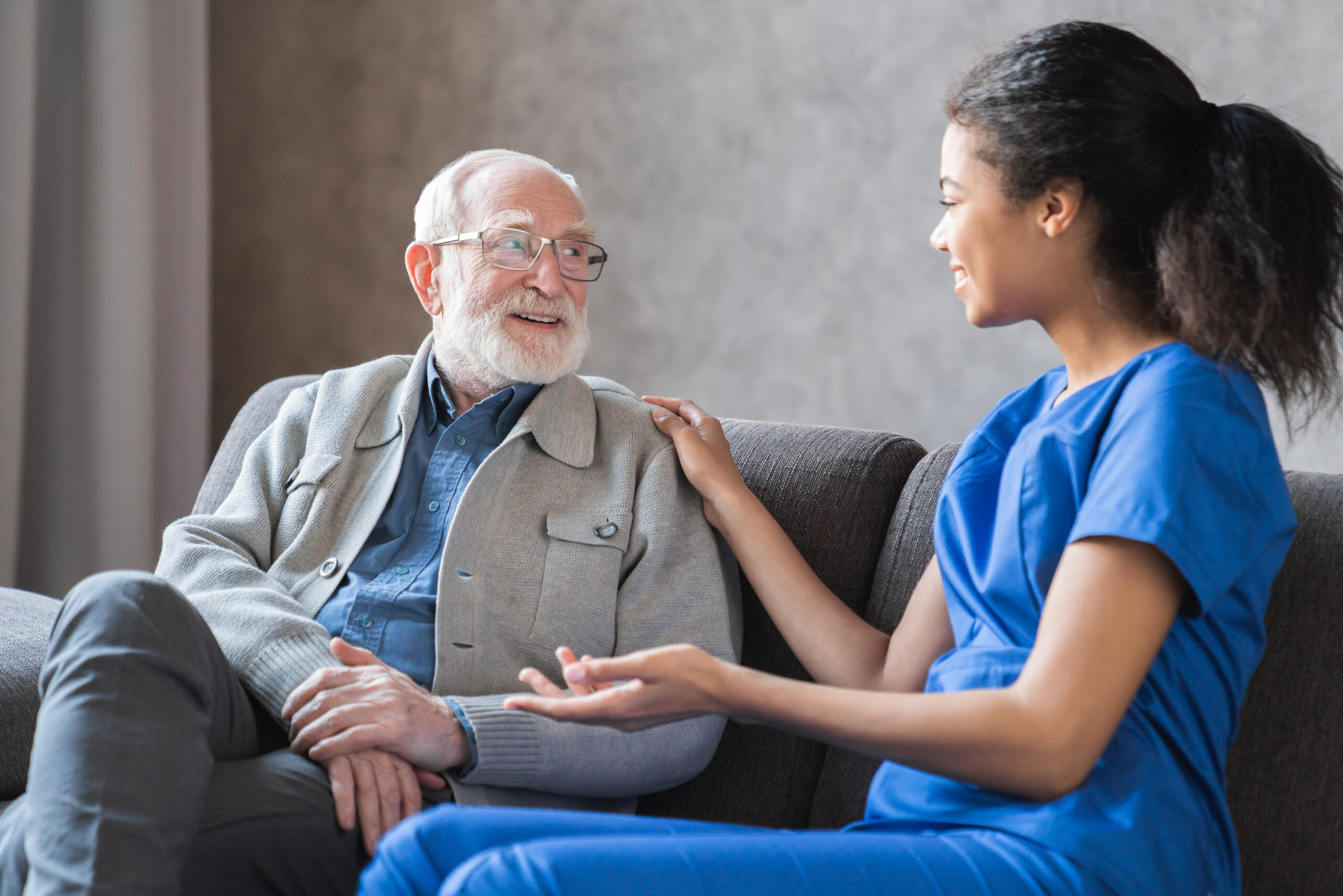 Compassionate caregiver in blue scrubs talking to a happy senior man on a couch, providing personalized senior care in Harrisburg, PA.