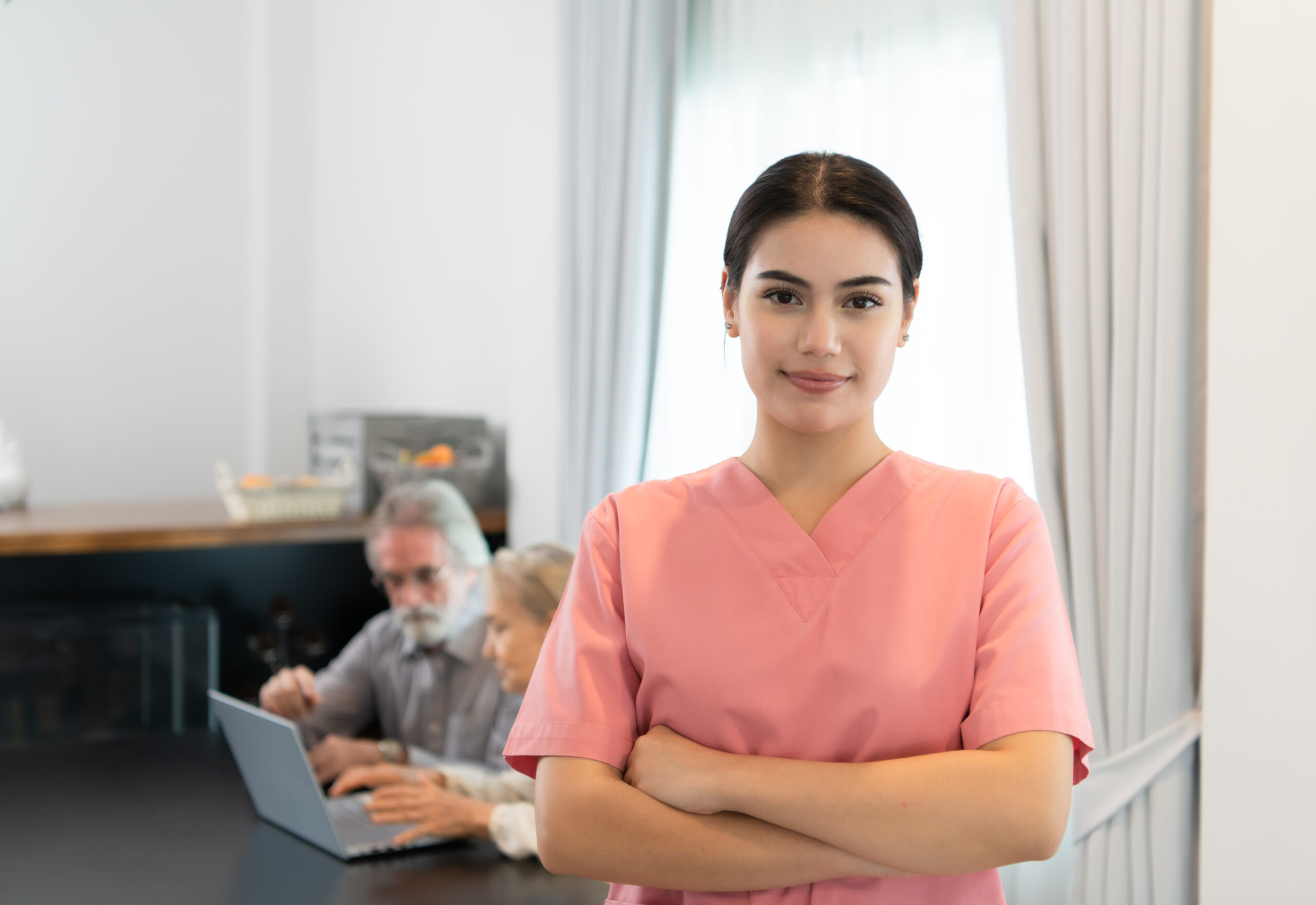 Confident young female caregiver in pink scrubs standing with arms crossed, smiling, with elderly clients in the background using a laptop. A representation of compassionate home care services.