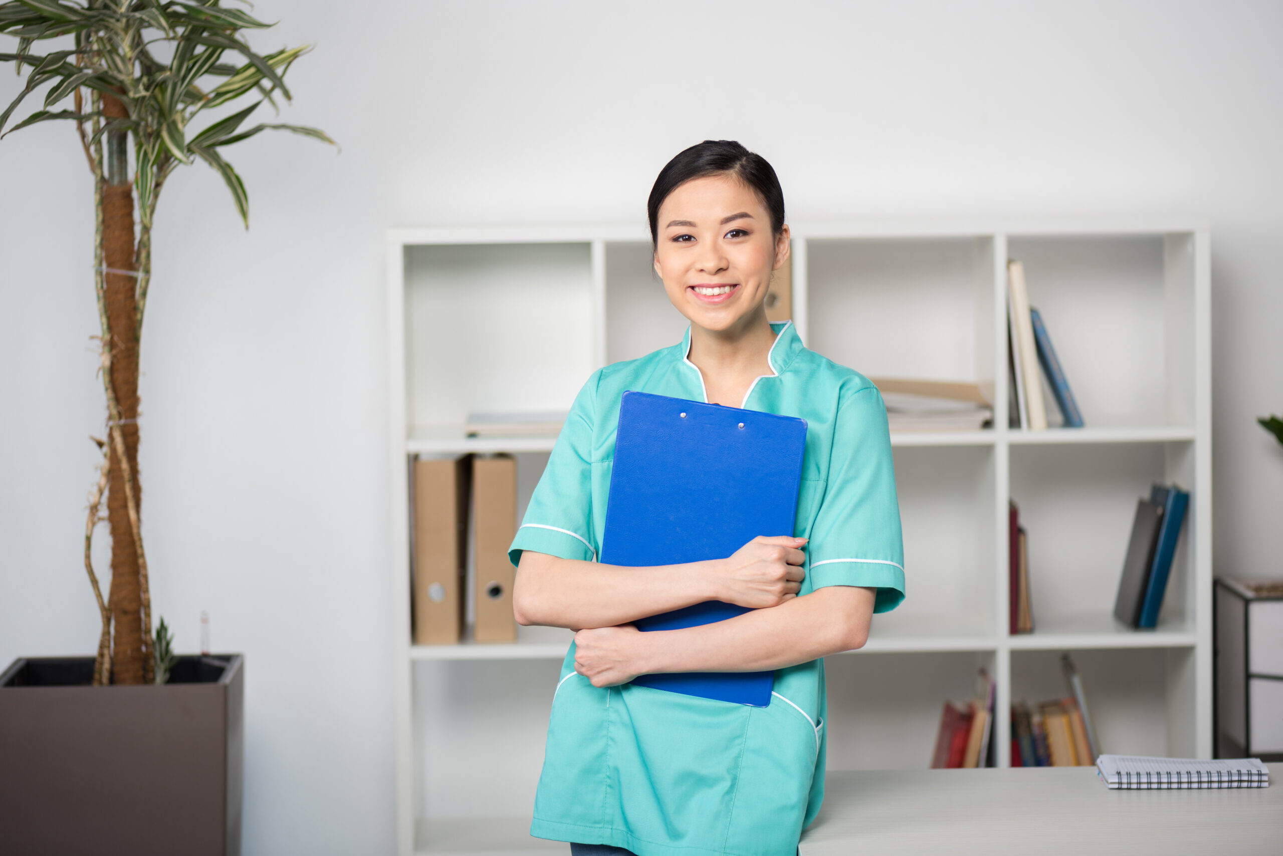 Smiling Asian female caregiver in a turquoise uniform holding a blue clipboard in a professional office setting. Representing AmeriBest Home Care, the best home care agency to work for in Philadelphia.